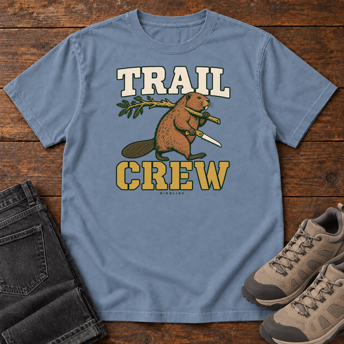 Blue Jean Trail Crew Foreman T-Shirt with a beaver carrying a leafy branch and saw beneath Trail Crew lettering; front flat lay on wood.