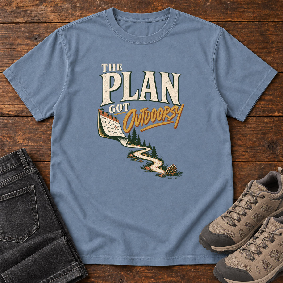 Blue Jean The Plan Got Outdoorsy T-Shirt with cream-and-gold lettering above a calendar turning into a winding trail, front flat lay on wood.