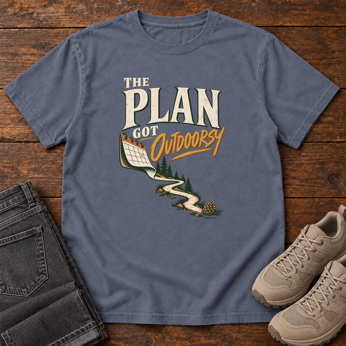 Denim The Plan Got Outdoorsy T-Shirt with cream-and-gold lettering above a calendar turning into a winding trail, front flat lay on wood.