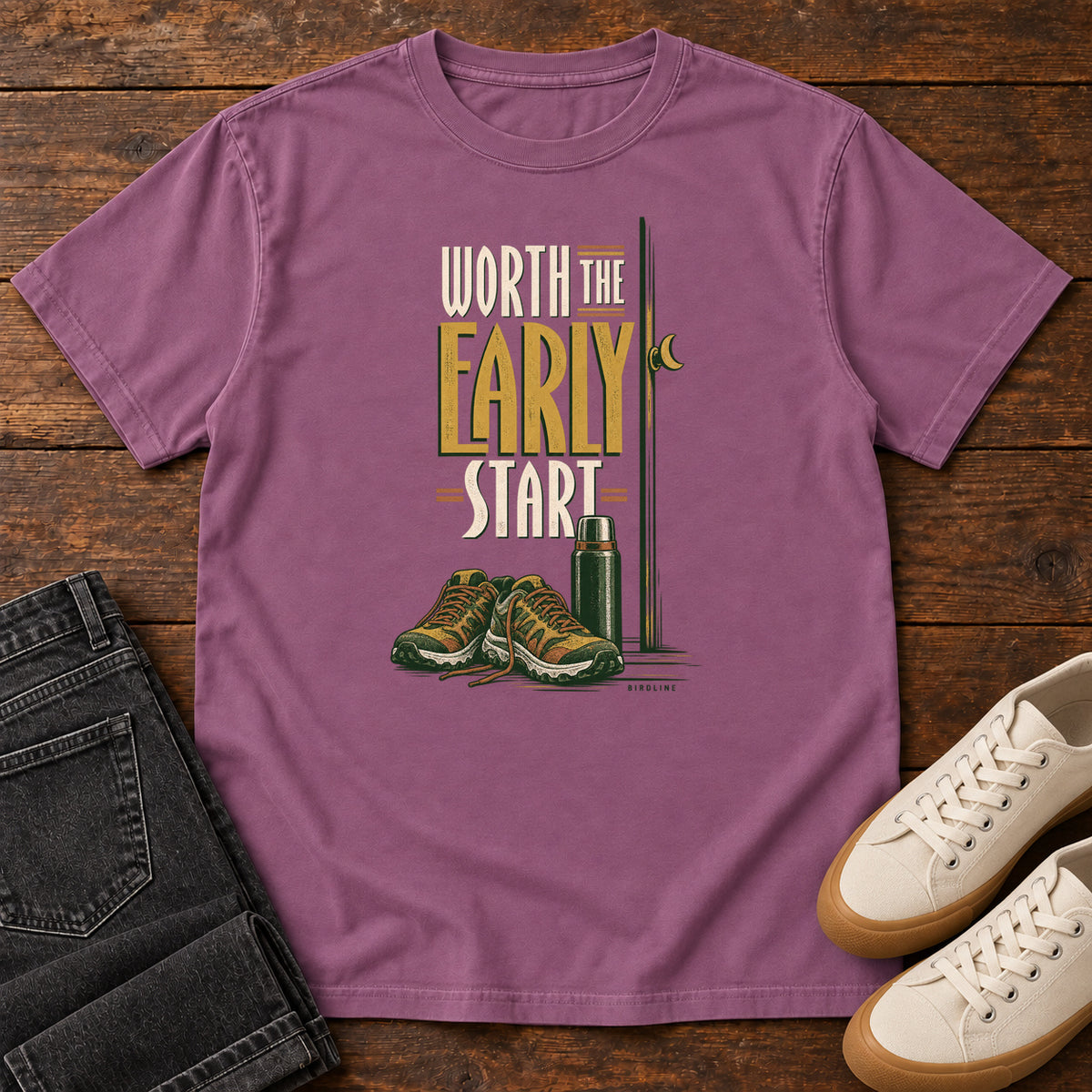 Berry Worth the Early Start T-Shirt with hiking shoes, a thermos, a walking pole and a crescent moon; front flat lay on wood.
