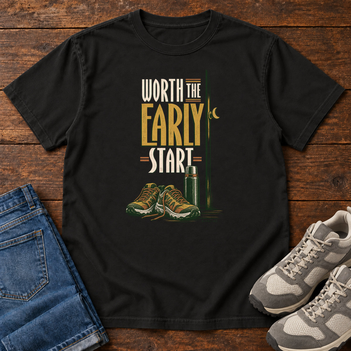 Black Worth the Early Start T-Shirt with hiking shoes, a thermos, a walking pole and a crescent moon; front flat lay on wood.
