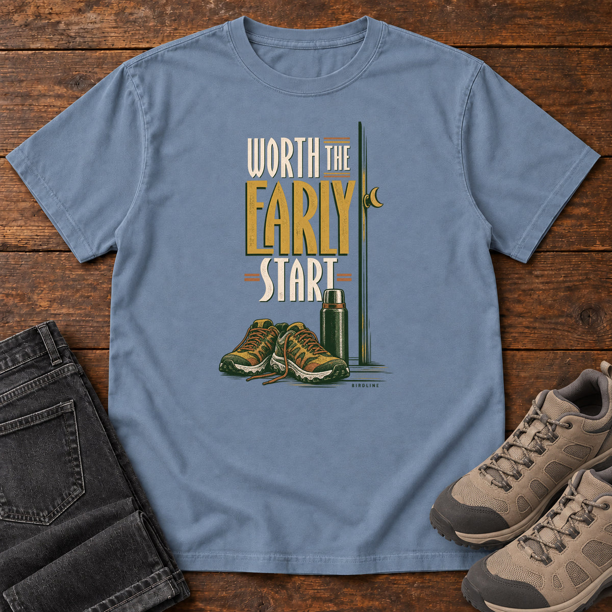 Blue Jean Worth the Early Start T-Shirt with hiking shoes, a thermos, a walking pole and a crescent moon; front flat lay on wood.