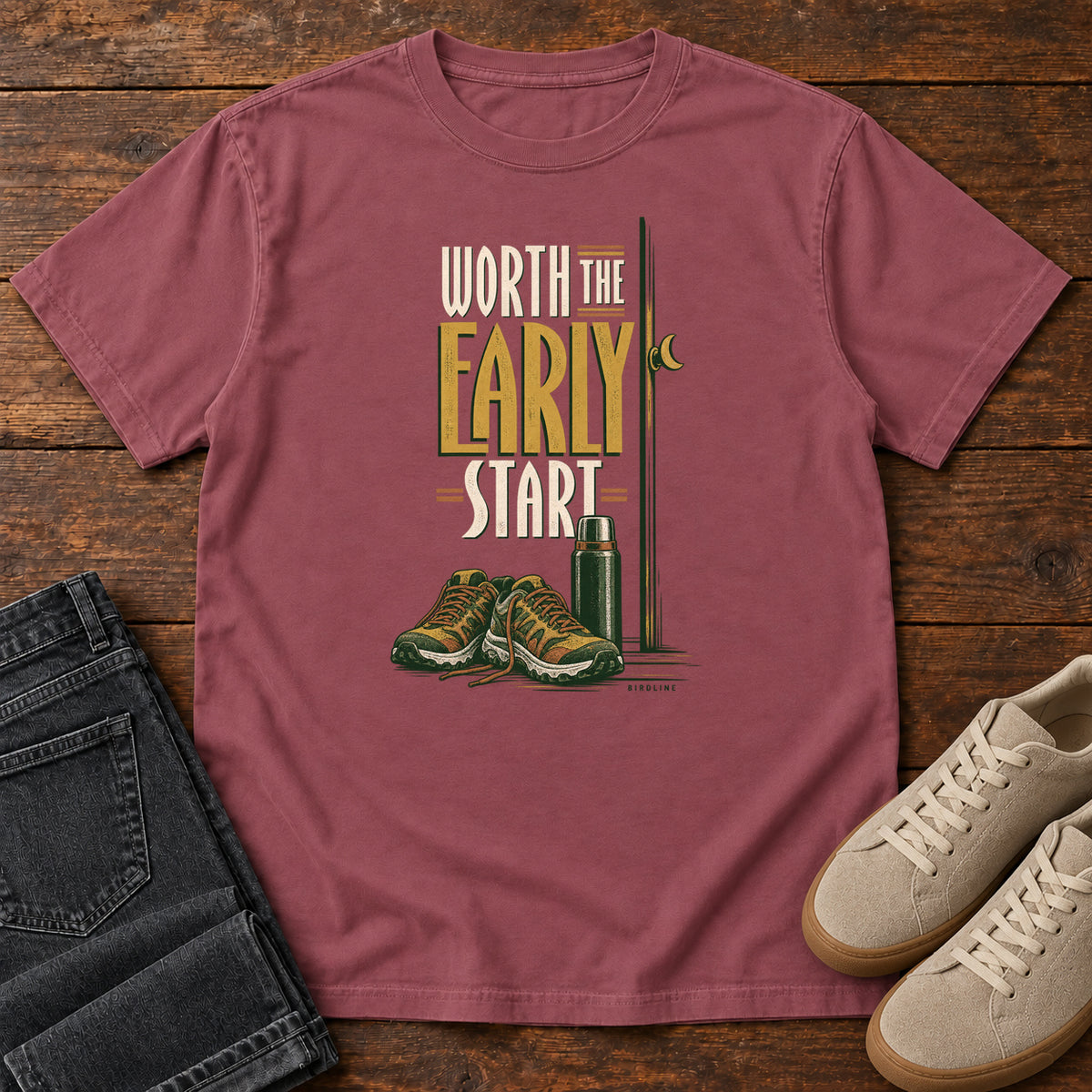 Brick Worth the Early Start T-Shirt with hiking shoes, a thermos, a walking pole and a crescent moon; front flat lay on wood.