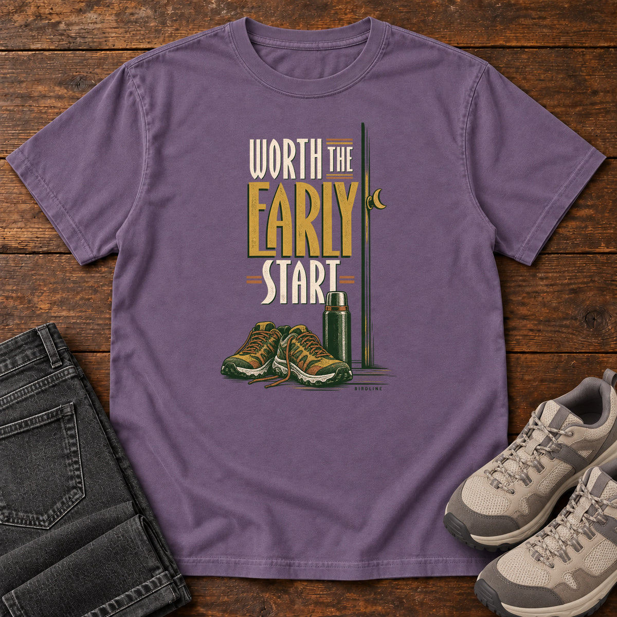 Grape Worth the Early Start T-Shirt with hiking shoes, a thermos, a walking pole and a crescent moon; front flat lay on wood.