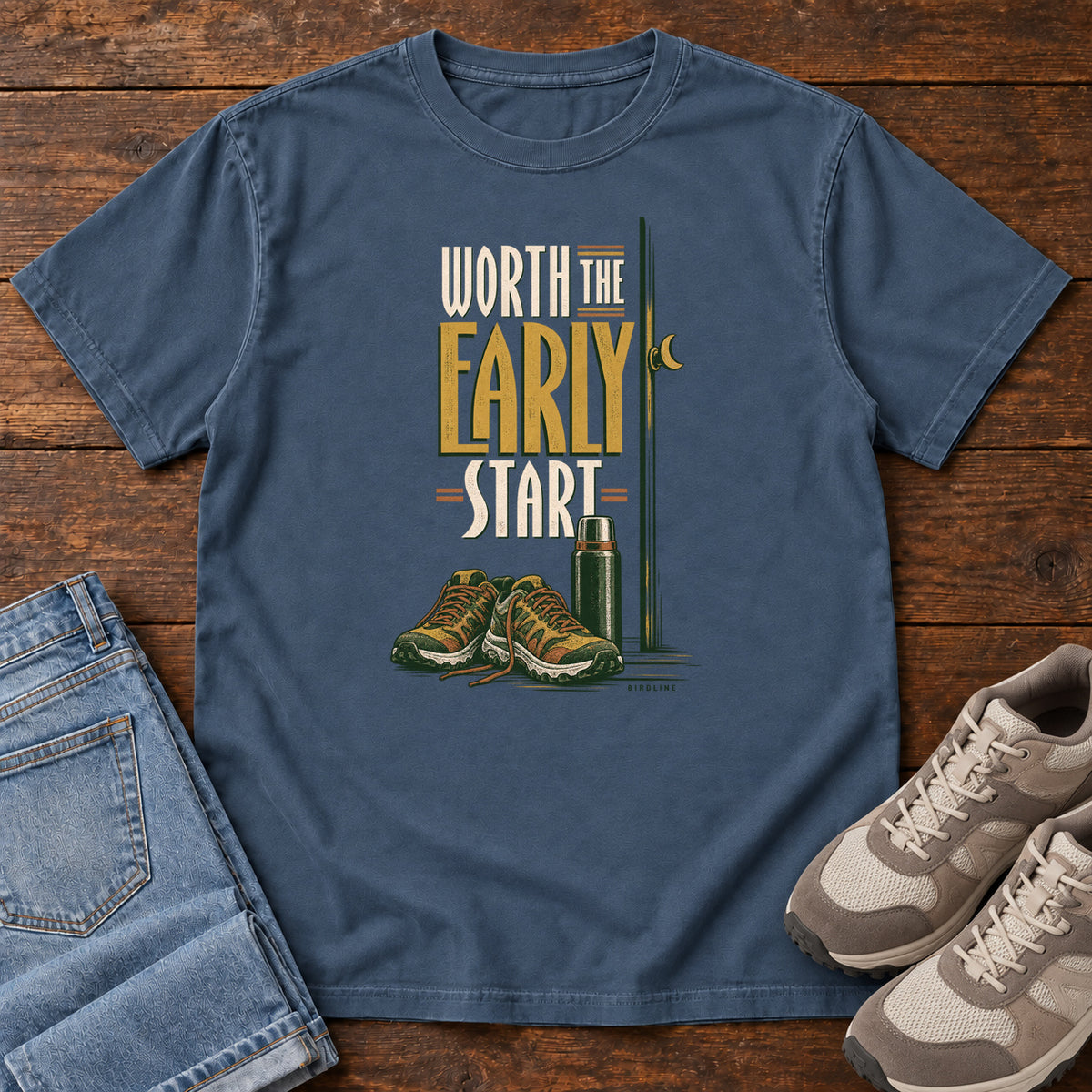 Midnight Worth the Early Start T-Shirt with hiking shoes, a thermos, a walking pole and a crescent moon; front flat lay on wood.