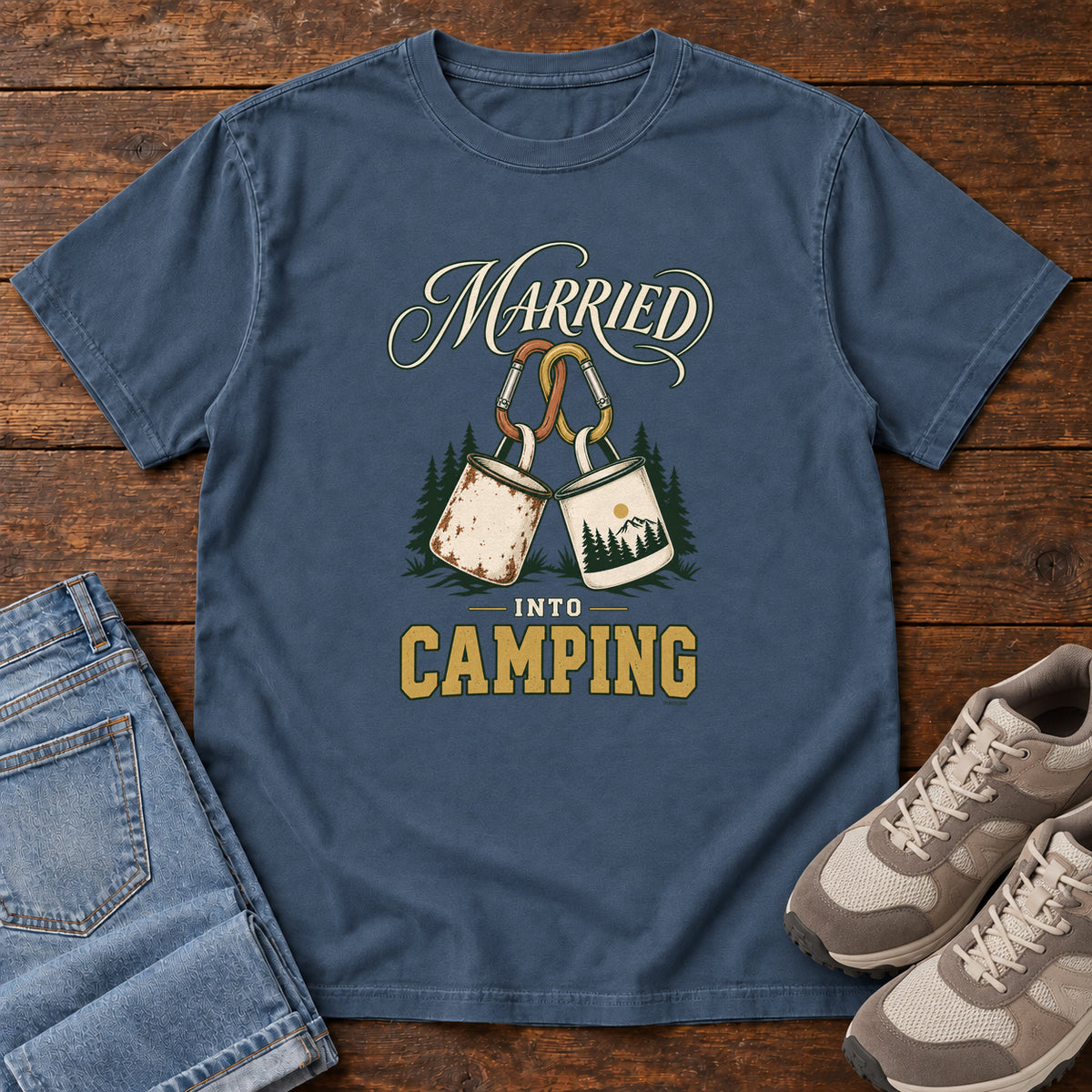 Midnight Married into Camping T-Shirt with two camp mugs hanging from carabiners between cream and gold lettering, front flat lay on wood.