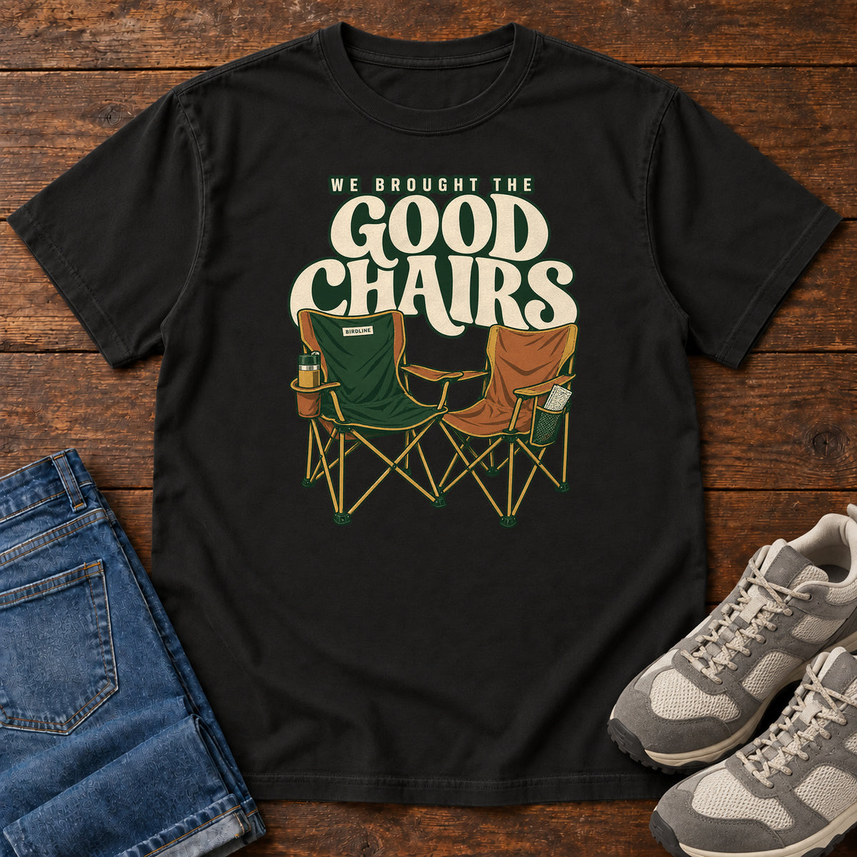 We Brought the Good Chairs T-Shirt in Black, front laid flat on wood, featuring green and brown folding camp chairs beneath large cream slogan lettering.