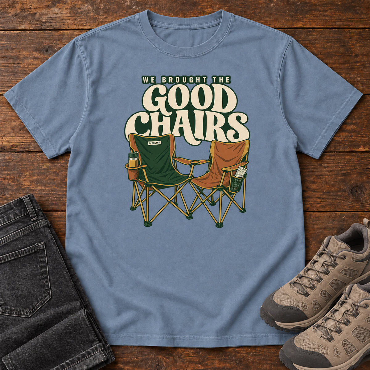 We Brought the Good Chairs T-Shirt in Blue Jean, front laid flat on wood, featuring green and brown folding camp chairs beneath large cream slogan lettering.