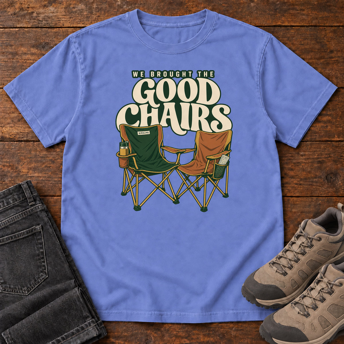 We Brought the Good Chairs T-Shirt in Flo Blue, front laid flat on wood, featuring green and brown folding camp chairs beneath large cream slogan lettering.