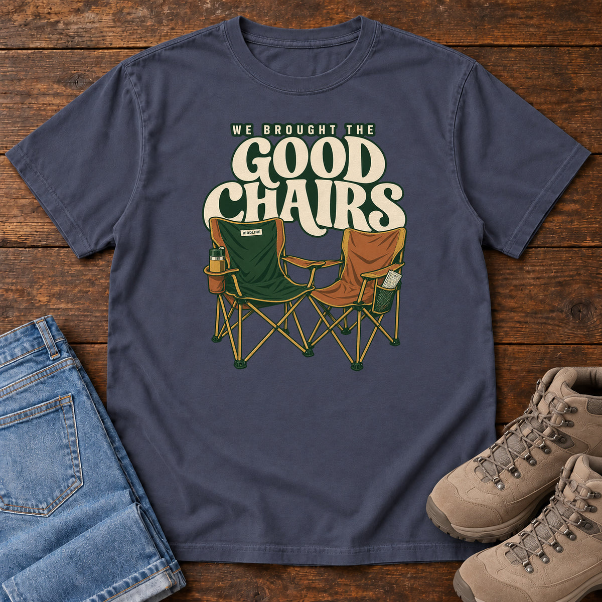 We Brought the Good Chairs T-Shirt in Navy, front laid flat on wood, featuring green and brown folding camp chairs beneath large cream slogan lettering.