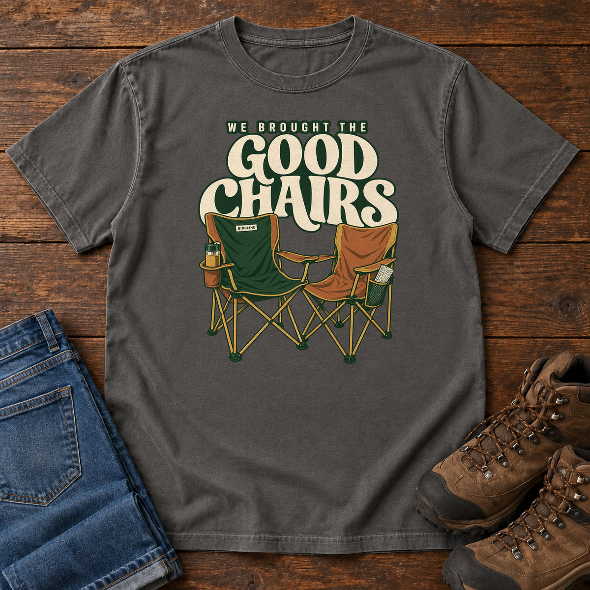 We Brought the Good Chairs T-Shirt in Pepper, front laid flat on wood, featuring green and brown folding camp chairs beneath large cream slogan lettering.