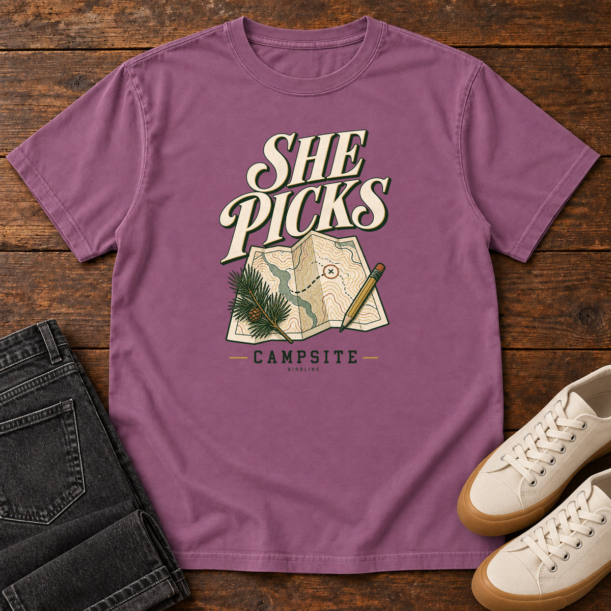 Berry She Picks the Campsite T-Shirt with cream lettering, a folded map, pencil and evergreen sprig, front flat lay on wood.