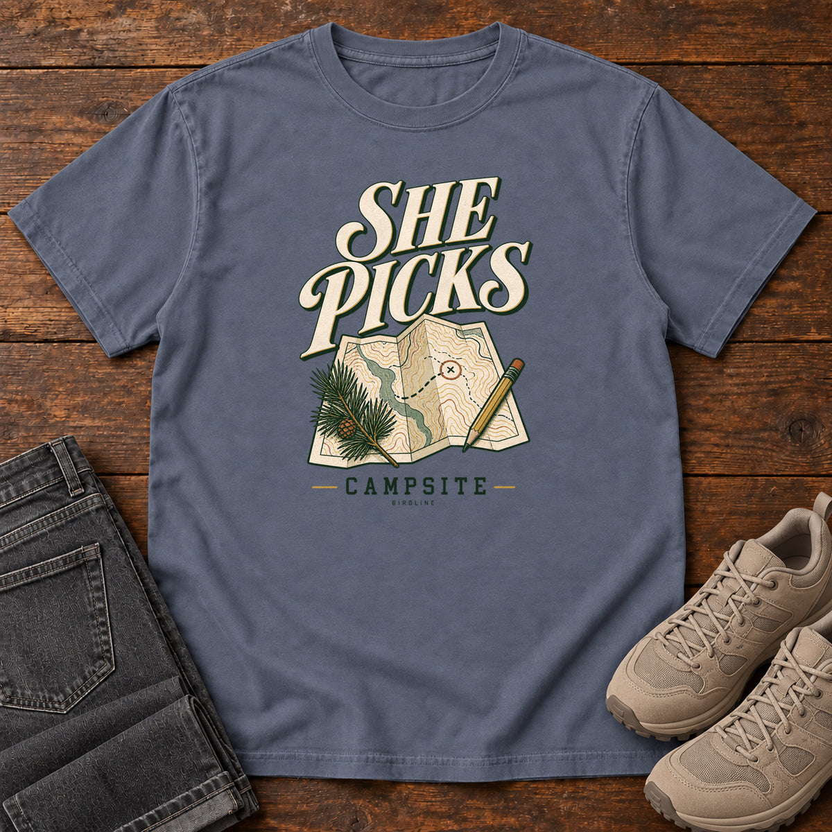 Denim She Picks the Campsite T-Shirt with cream lettering, a folded map, pencil and evergreen sprig, front flat lay on wood.