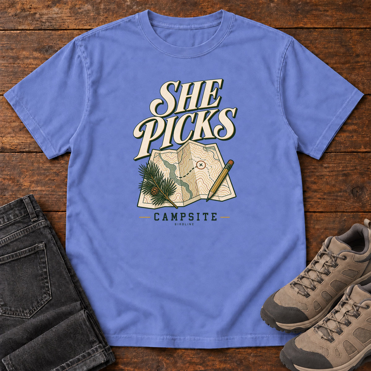 Flo Blue She Picks the Campsite T-Shirt with cream lettering, a folded map, pencil and evergreen sprig, front flat lay on wood.