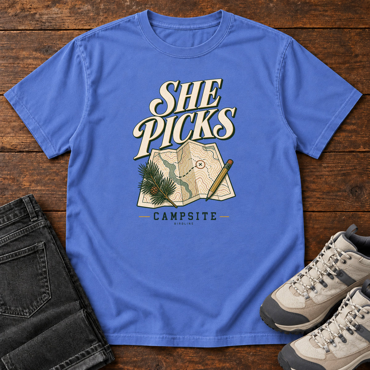 Mystic Blue She Picks the Campsite T-Shirt with cream lettering, a folded map, pencil and evergreen sprig, front flat lay on wood.