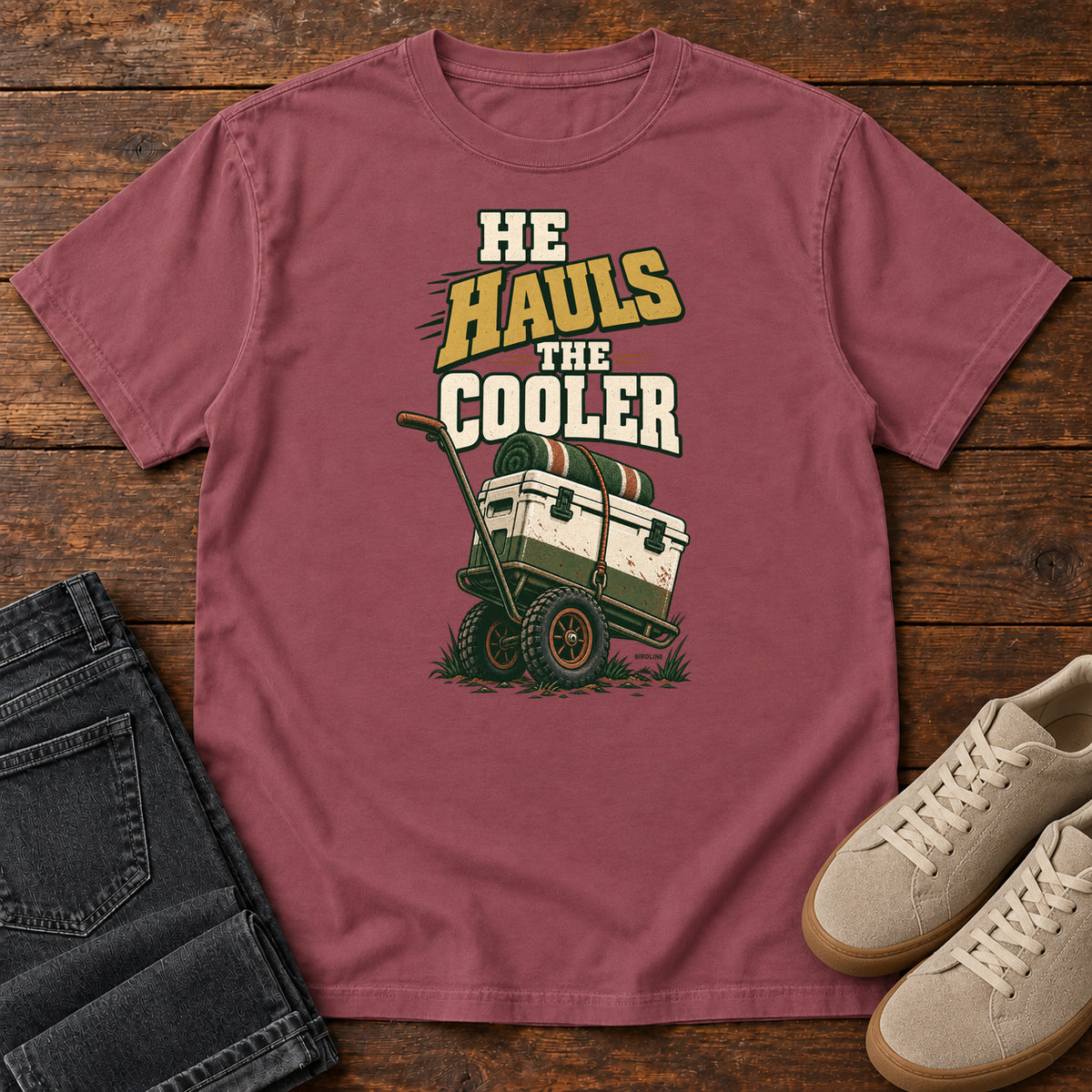 Brick He Hauls the Cooler T-Shirt with a cooler on a two-wheeled cart beneath He Hauls the Cooler lettering; front flat lay on wood.