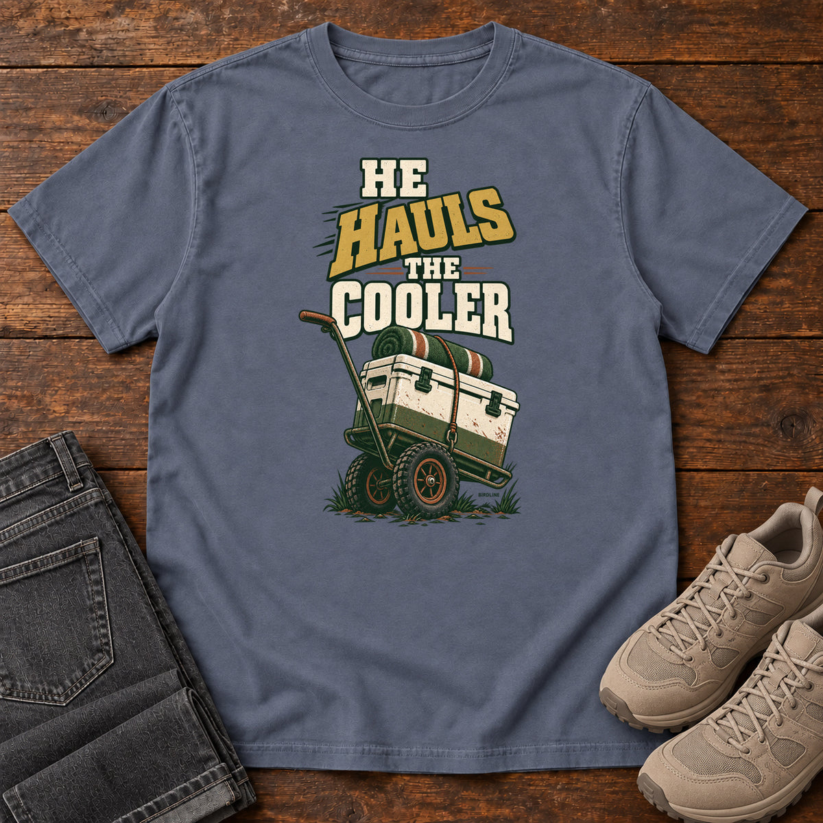 Denim He Hauls the Cooler T-Shirt with a cooler on a two-wheeled cart beneath He Hauls the Cooler lettering; front flat lay on wood.