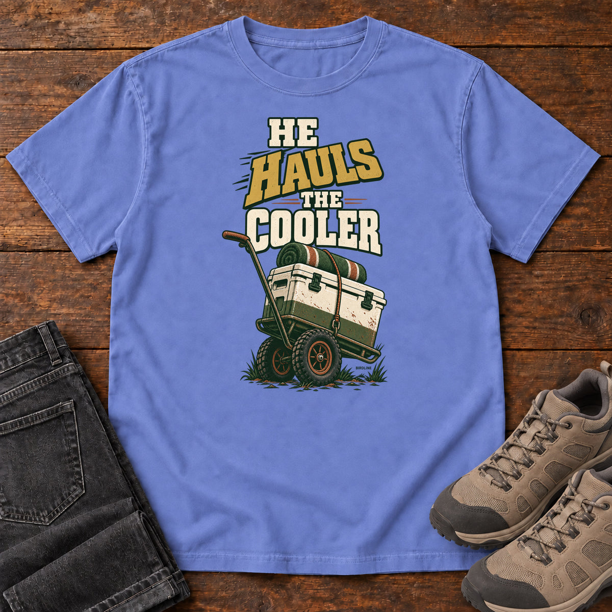 Flo Blue He Hauls the Cooler T-Shirt with a cooler on a two-wheeled cart beneath He Hauls the Cooler lettering; front flat lay on wood.
