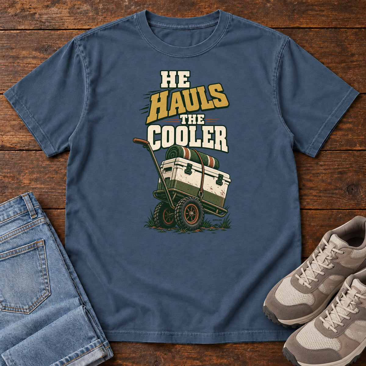 Midnight He Hauls the Cooler T-Shirt with a cooler on a two-wheeled cart beneath He Hauls the Cooler lettering; front flat lay on wood.