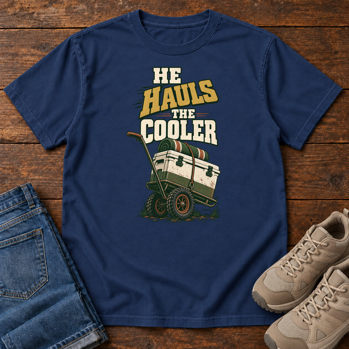 True Navy He Hauls the Cooler T-Shirt with a cooler on a two-wheeled cart beneath He Hauls the Cooler lettering; front flat lay on wood.