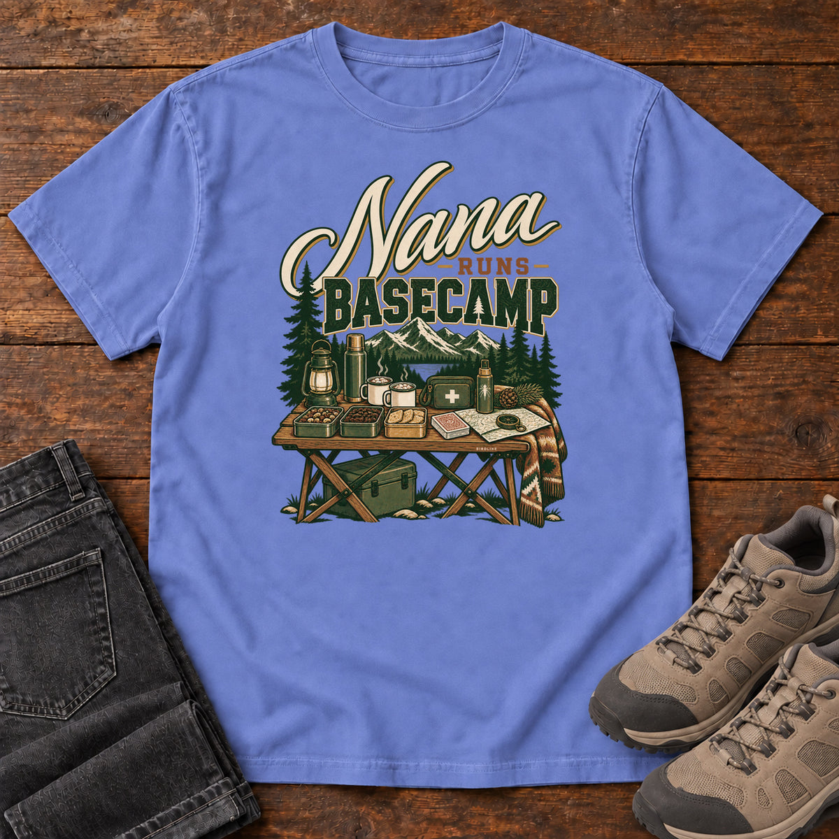 Flo Blue Nana Runs Basecamp T-Shirt with a camp table with mugs, lantern, snacks and supplies beneath mountains; front flat lay on wood.