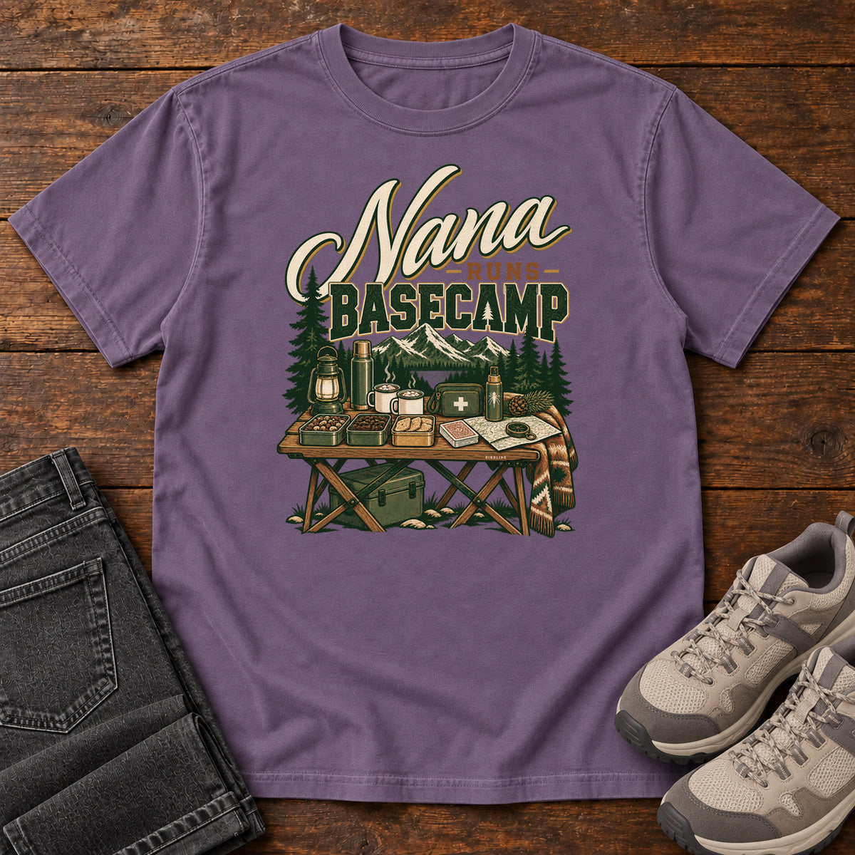 Grape Nana Runs Basecamp T-Shirt with a camp table with mugs, lantern, snacks and supplies beneath mountains; front flat lay on wood.