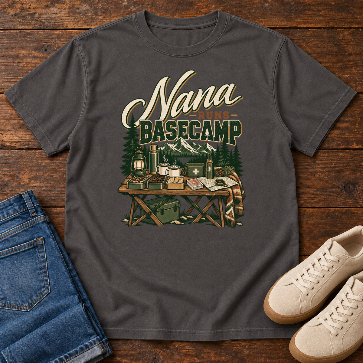 Graphite Nana Runs Basecamp T-Shirt with a camp table with mugs, lantern, snacks and supplies beneath mountains; front flat lay on wood.