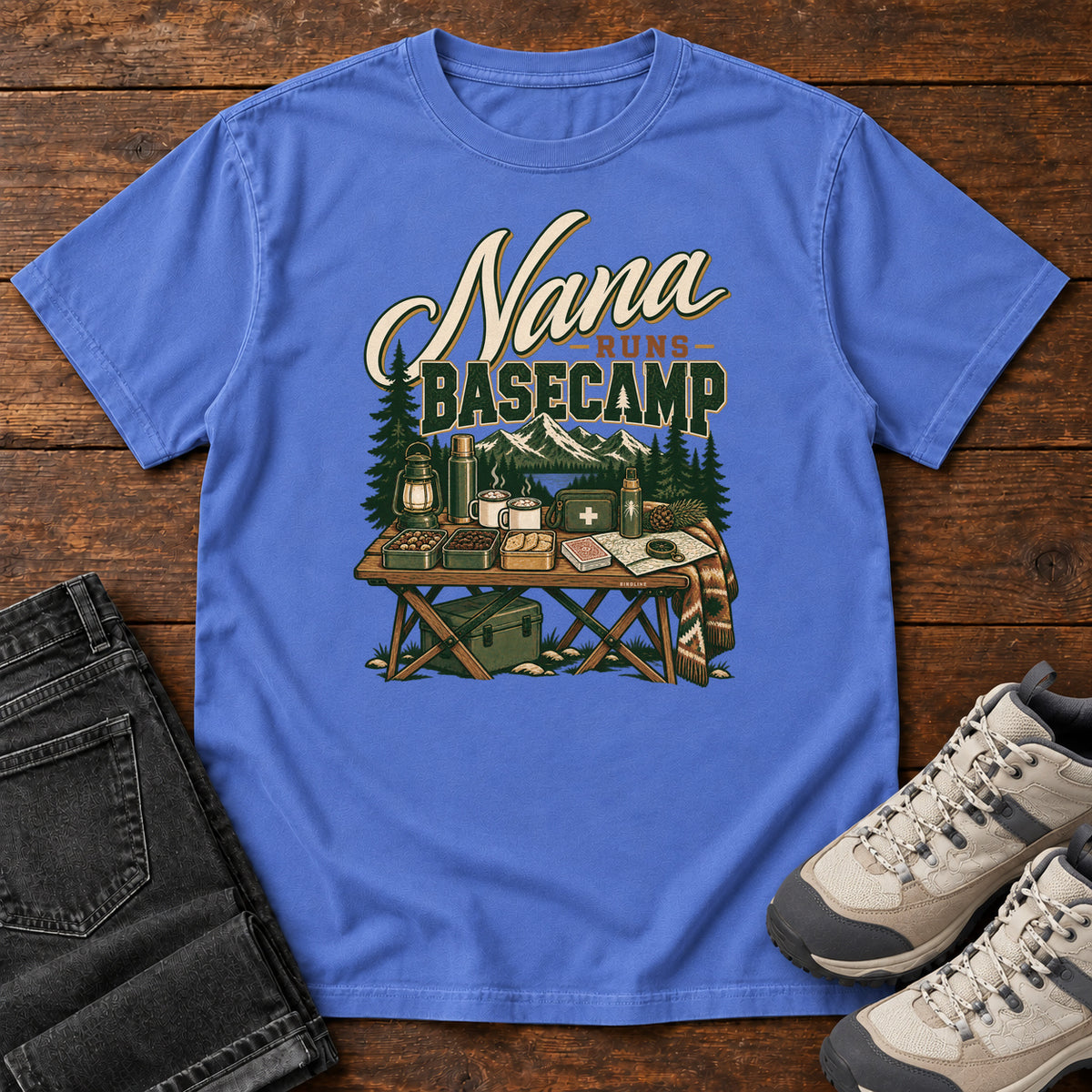 Mystic Blue Nana Runs Basecamp T-Shirt with a camp table with mugs, lantern, snacks and supplies beneath mountains; front flat lay on wood.