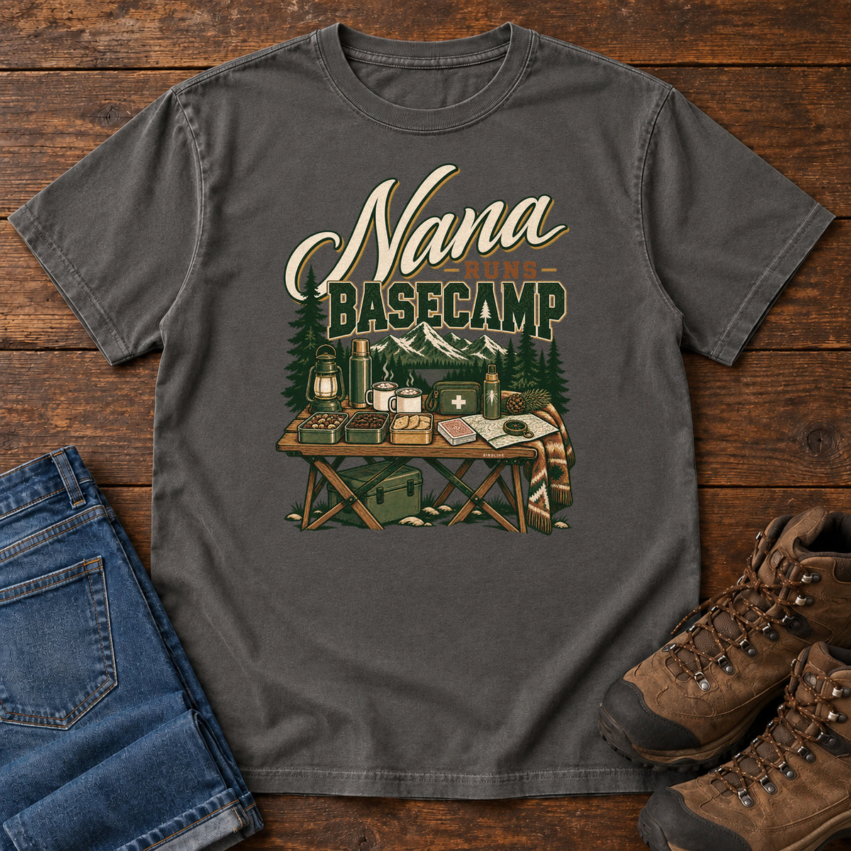 Pepper Nana Runs Basecamp T-Shirt with a camp table with mugs, lantern, snacks and supplies beneath mountains; front flat lay on wood.