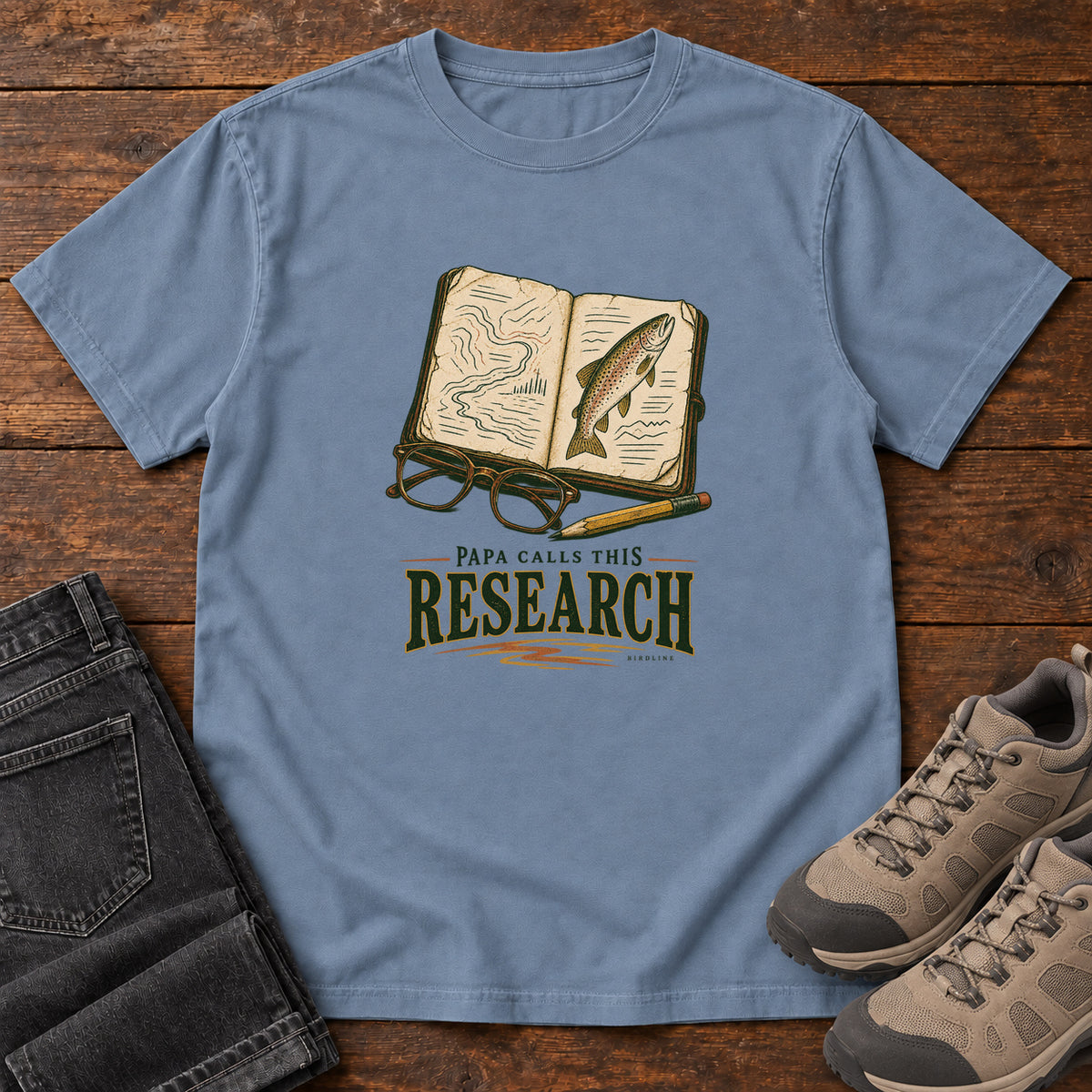 Blue Jean Papa Calls This Research T-Shirt with an open field notebook showing a map and fish, with glasses and a pencil, front flat lay on wood.