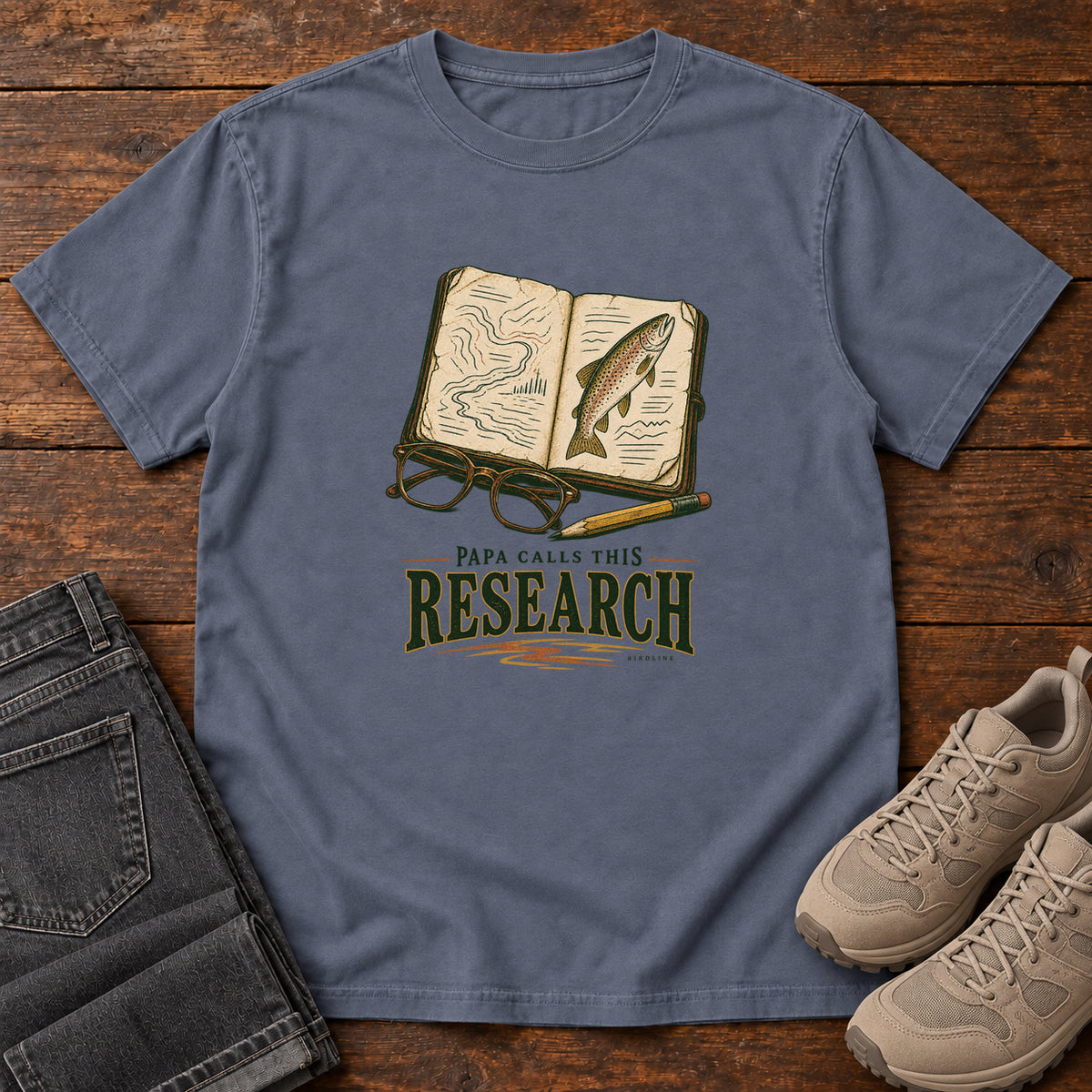Denim Papa Calls This Research T-Shirt with an open field notebook showing a map and fish, with glasses and a pencil, front flat lay on wood.