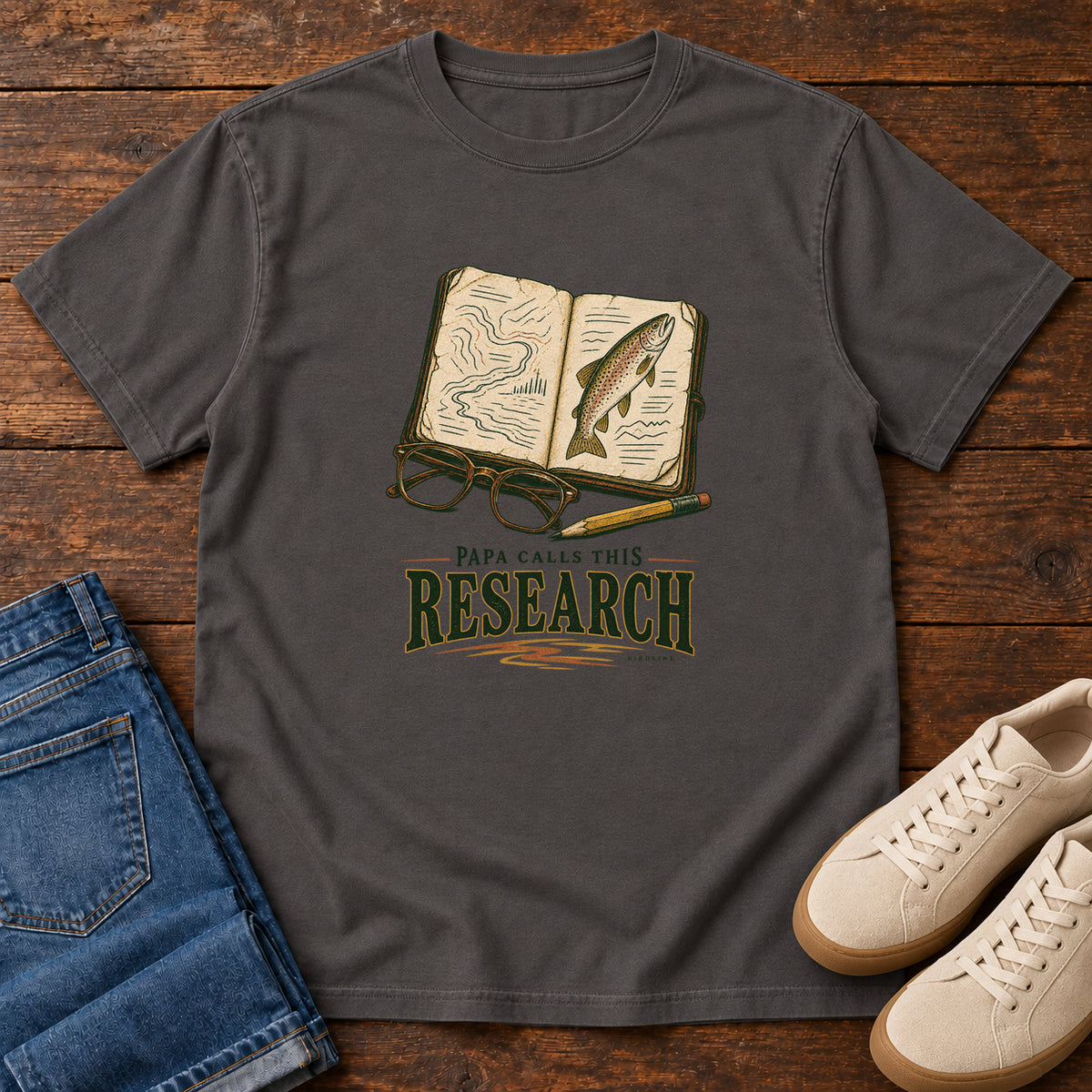 Graphite Papa Calls This Research T-Shirt with an open field notebook showing a map and fish, with glasses and a pencil, front flat lay on wood.