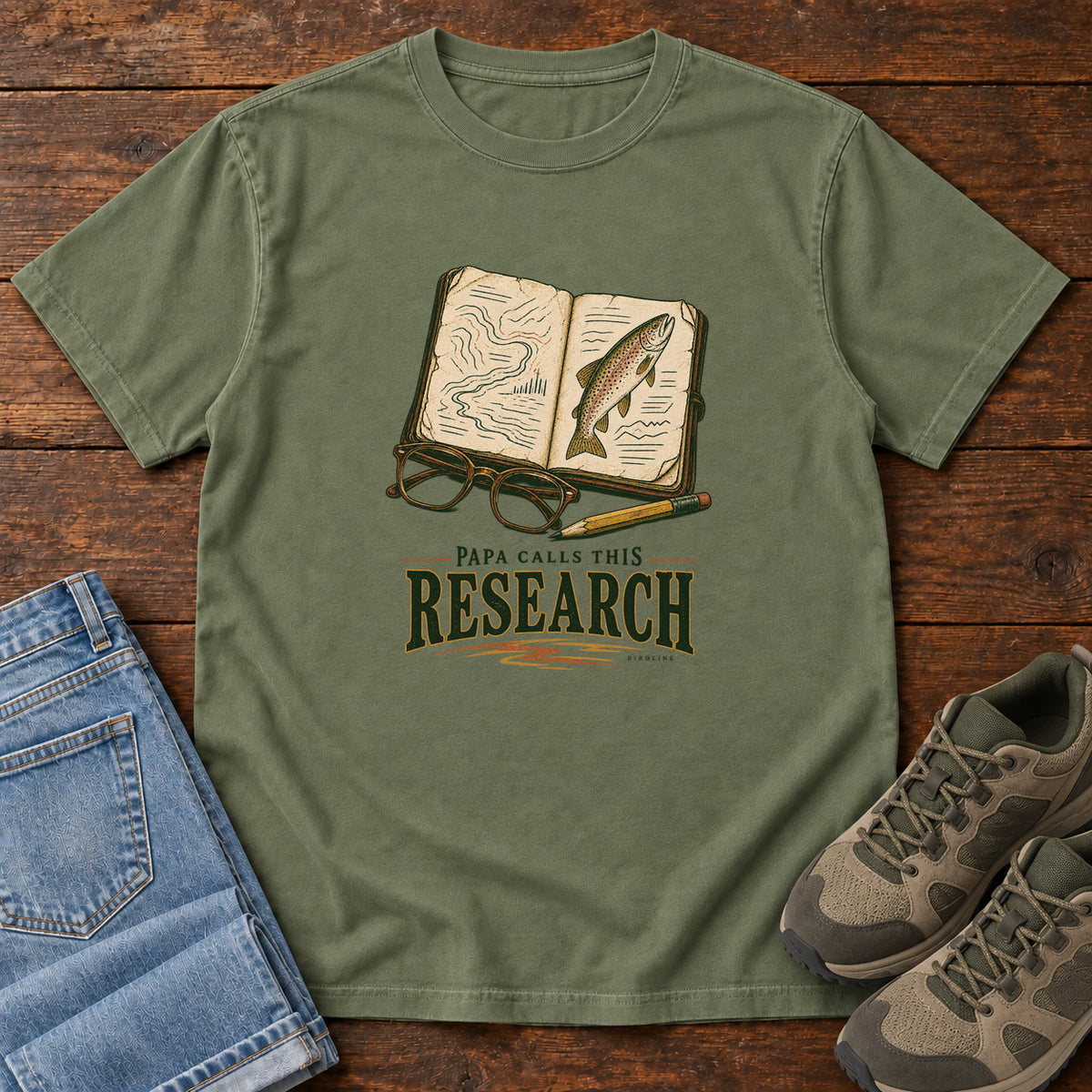 Moss Papa Calls This Research T-Shirt with an open field notebook showing a map and fish, with glasses and a pencil, front flat lay on wood.