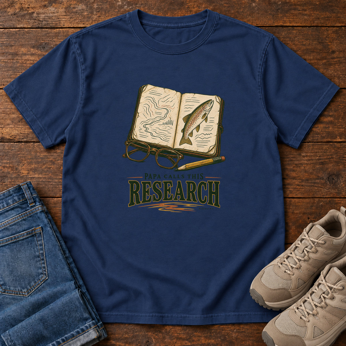 True Navy Papa Calls This Research T-Shirt with an open field notebook showing a map and fish, with glasses and a pencil, front flat lay on wood.