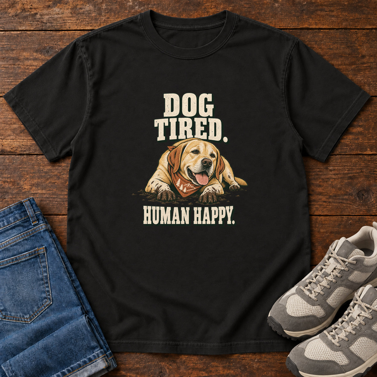 Dog Tired Human Happy T-Shirt in Black, front laid flat on wood, featuring a resting golden-brown dog wearing an orange bandana between cream slogan lettering.