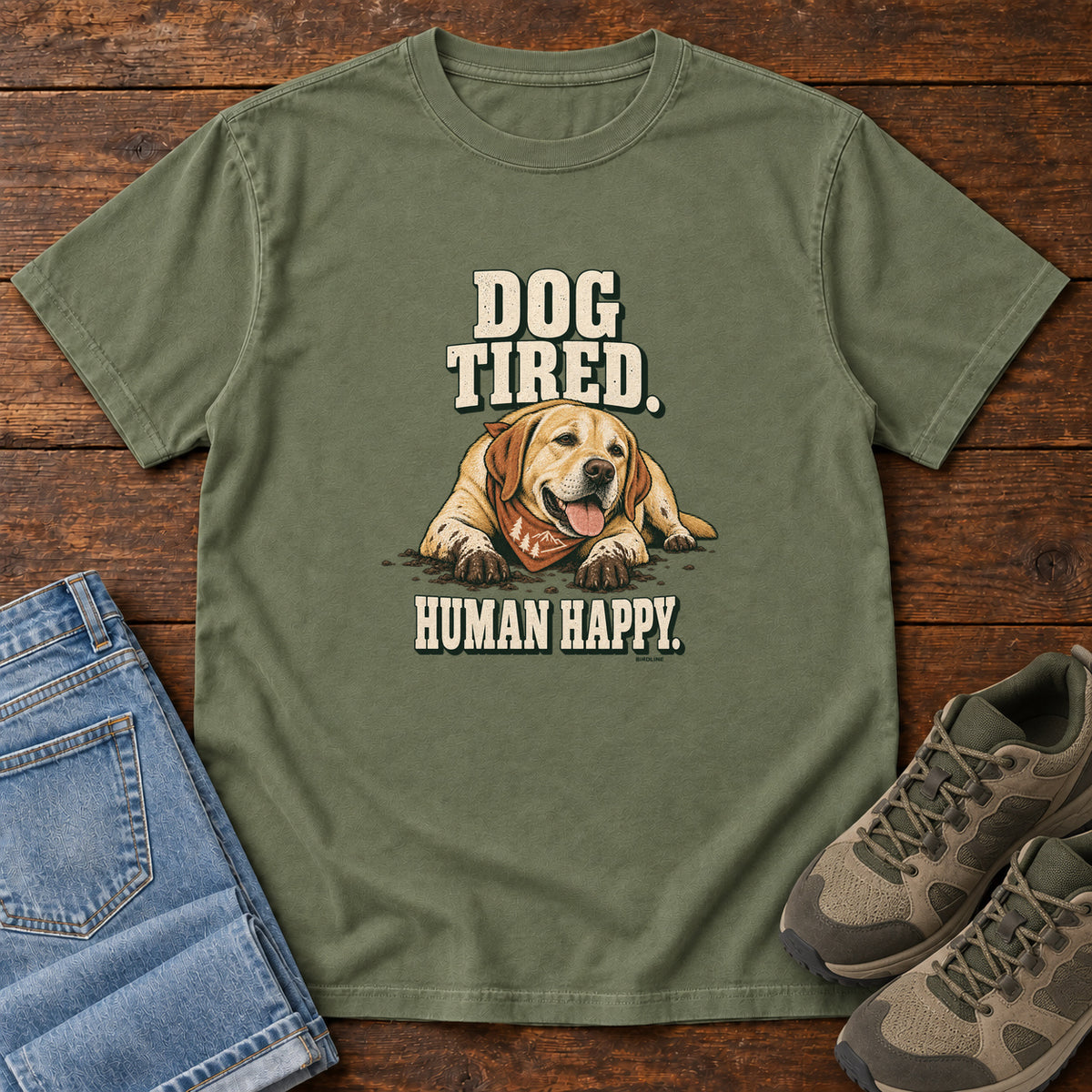 Dog Tired Human Happy T-Shirt in Moss, front laid flat on wood, featuring a resting golden-brown dog wearing an orange bandana between cream slogan lettering.