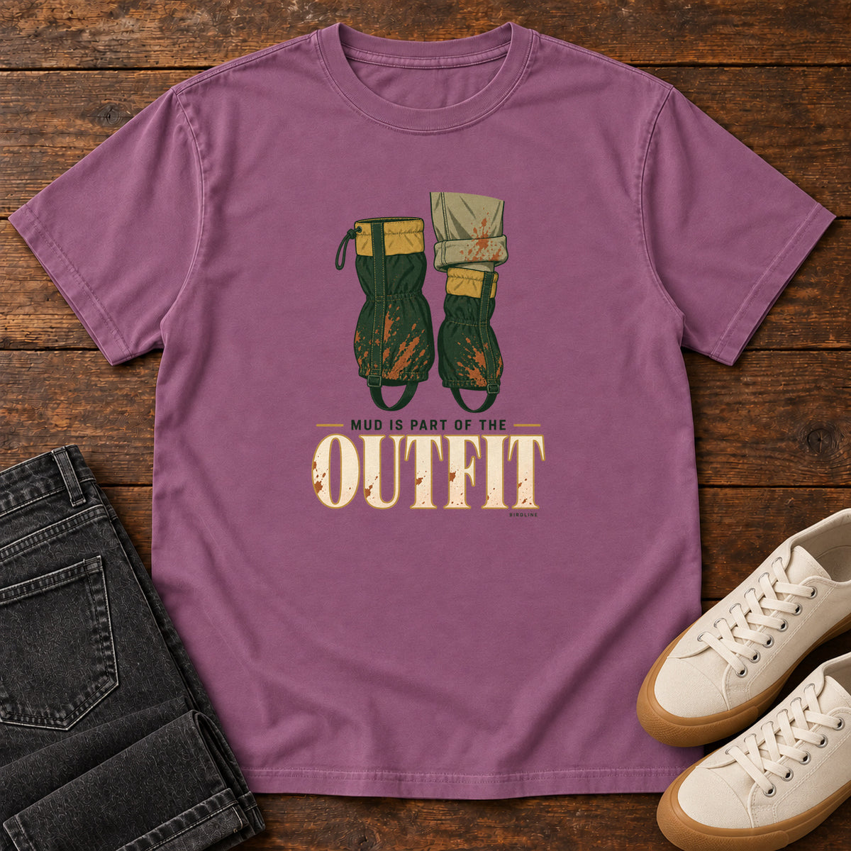 Berry Mud Is Part of the Outfit T-Shirt with mud-splashed green boots and cream lettering, front flat lay on wood.
