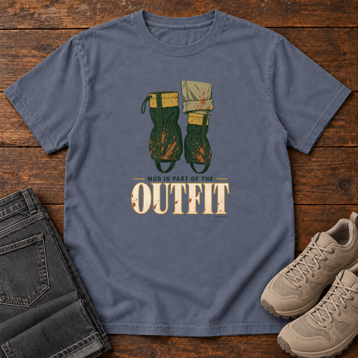Denim Mud Is Part of the Outfit T-Shirt with mud-splashed green boots and cream lettering, front flat lay on wood.