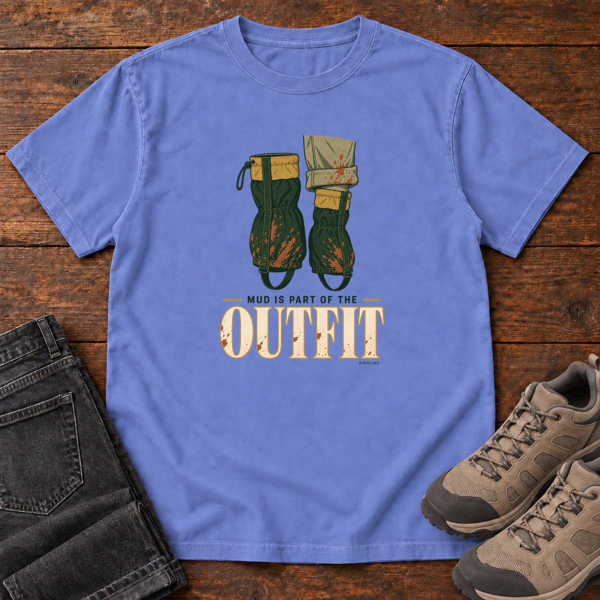Flo Blue Mud Is Part of the Outfit T-Shirt with mud-splashed green boots and cream lettering, front flat lay on wood.