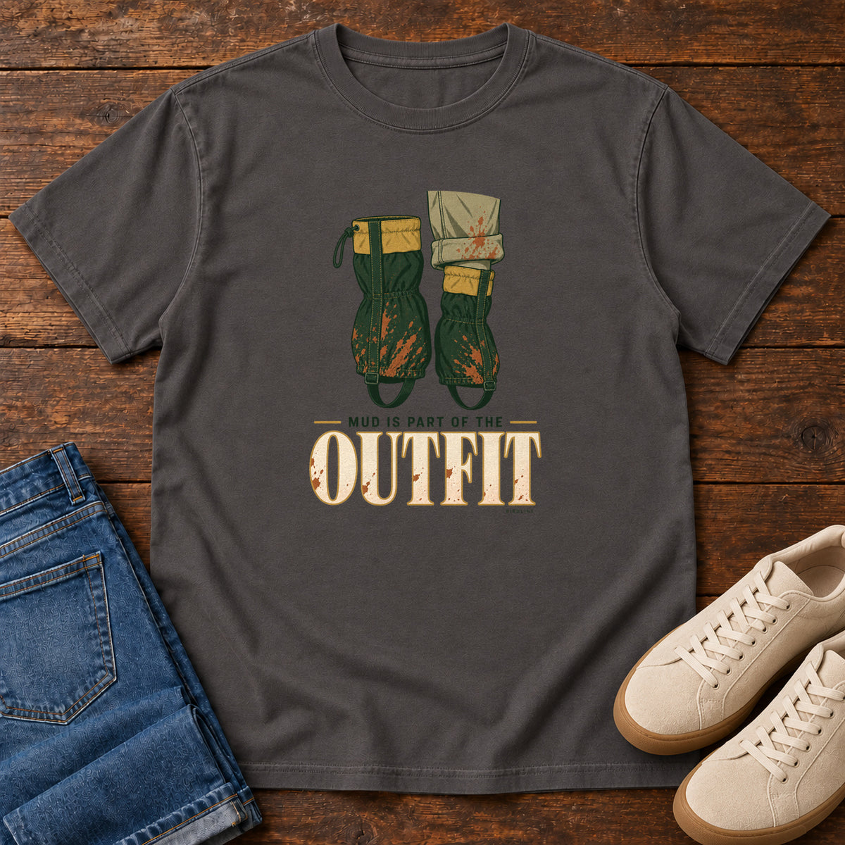 Graphite Mud Is Part of the Outfit T-Shirt with mud-splashed green boots and cream lettering, front flat lay on wood.
