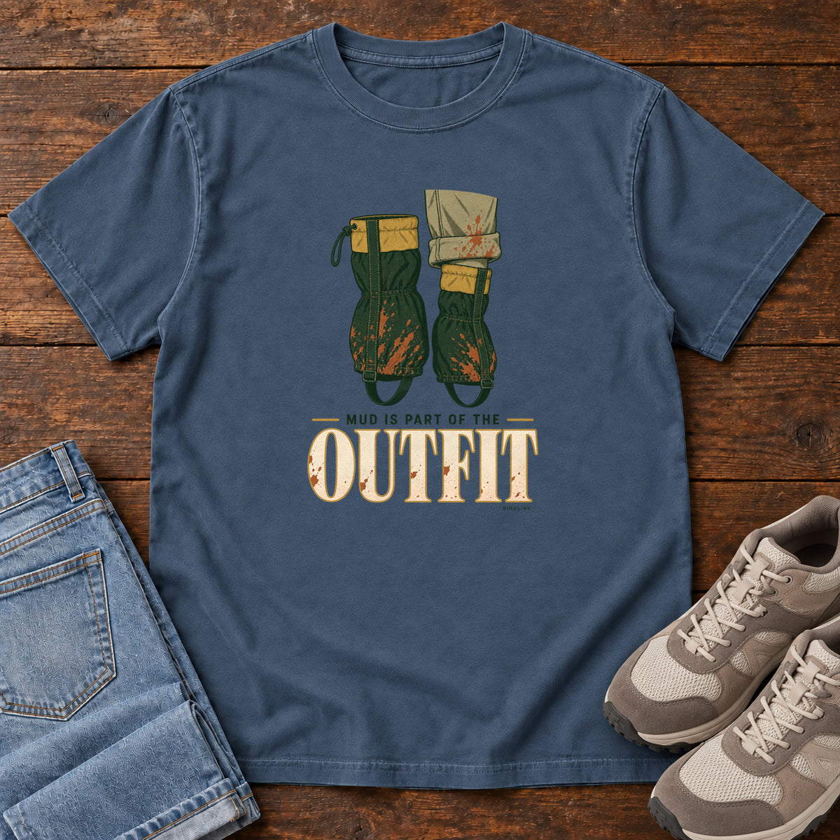 Midnight Mud Is Part of the Outfit T-Shirt with mud-splashed green boots and cream lettering, front flat lay on wood.