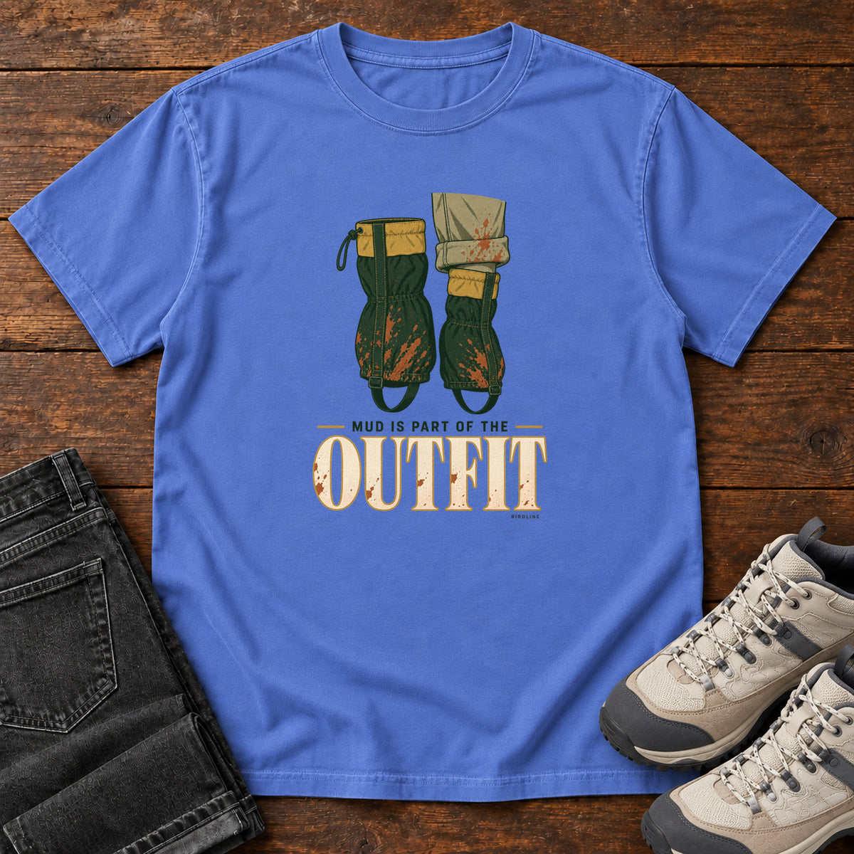 Mystic Blue Mud Is Part of the Outfit T-Shirt with mud-splashed green boots and cream lettering, front flat lay on wood.