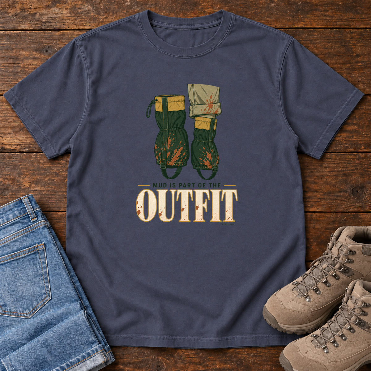 Navy Mud Is Part of the Outfit T-Shirt with mud-splashed green boots and cream lettering, front flat lay on wood.