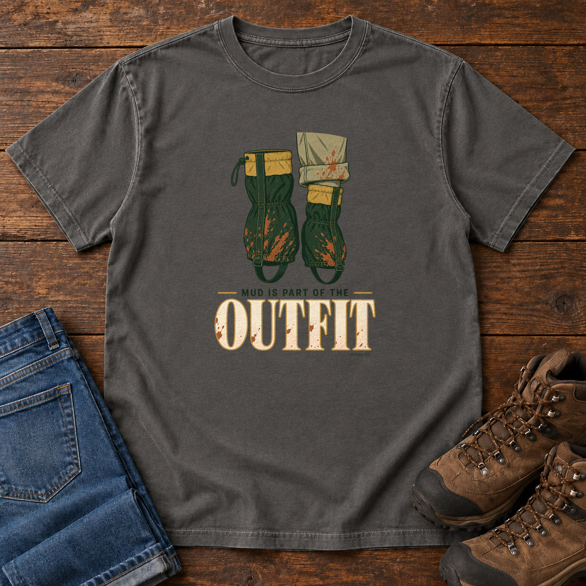 Pepper Mud Is Part of the Outfit T-Shirt with mud-splashed green boots and cream lettering, front flat lay on wood.