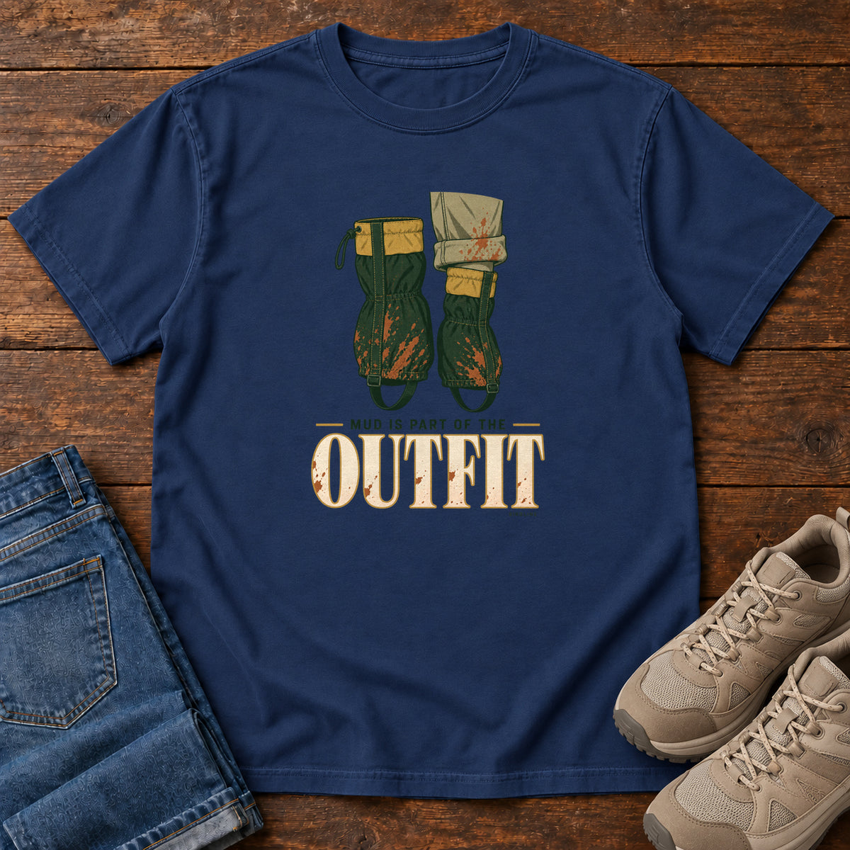 True Navy Mud Is Part of the Outfit T-Shirt with mud-splashed green boots and cream lettering, front flat lay on wood.