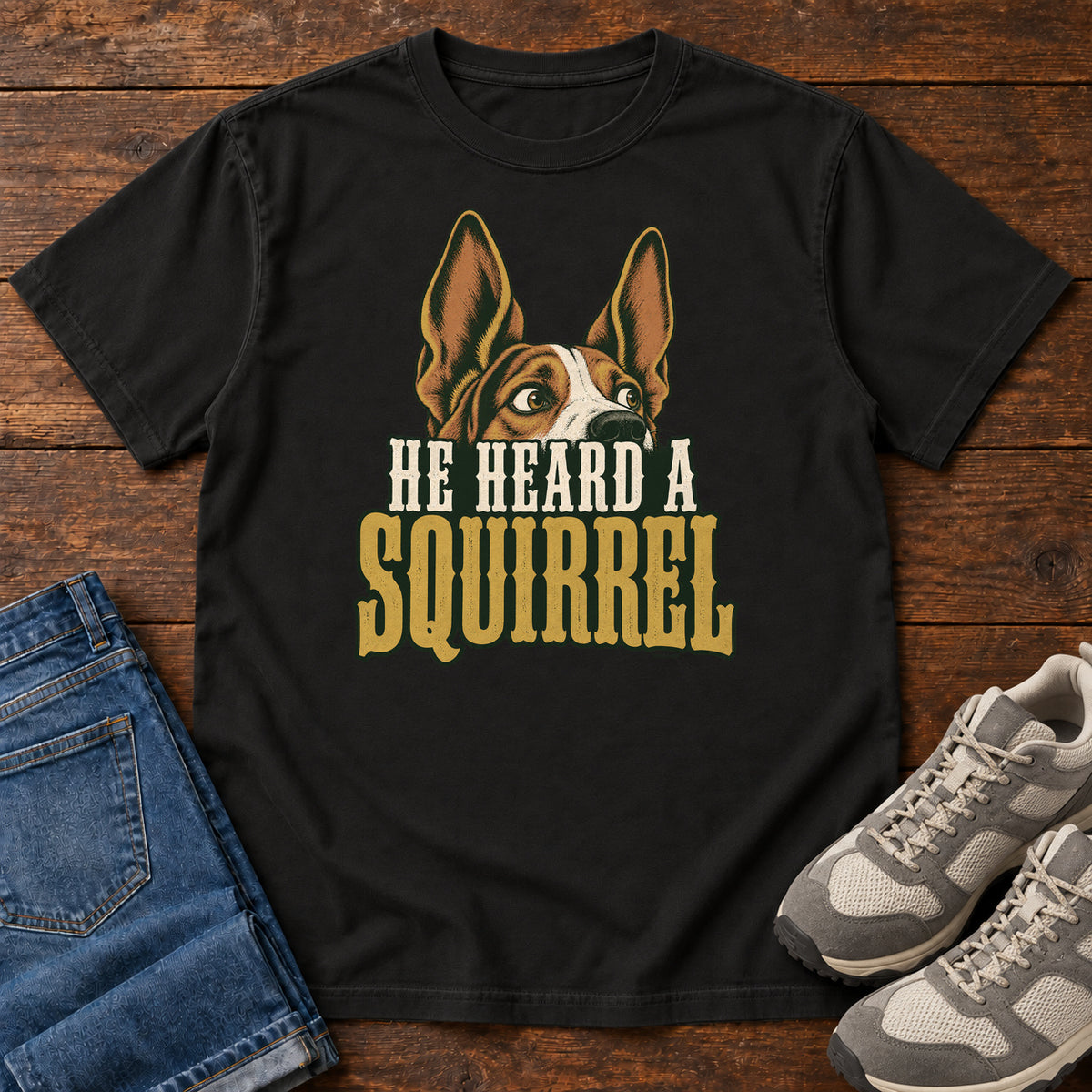 Black He Heard a Squirrel T-Shirt with an alert dog with upright ears above He Heard a Squirrel lettering; front flat lay on wood.