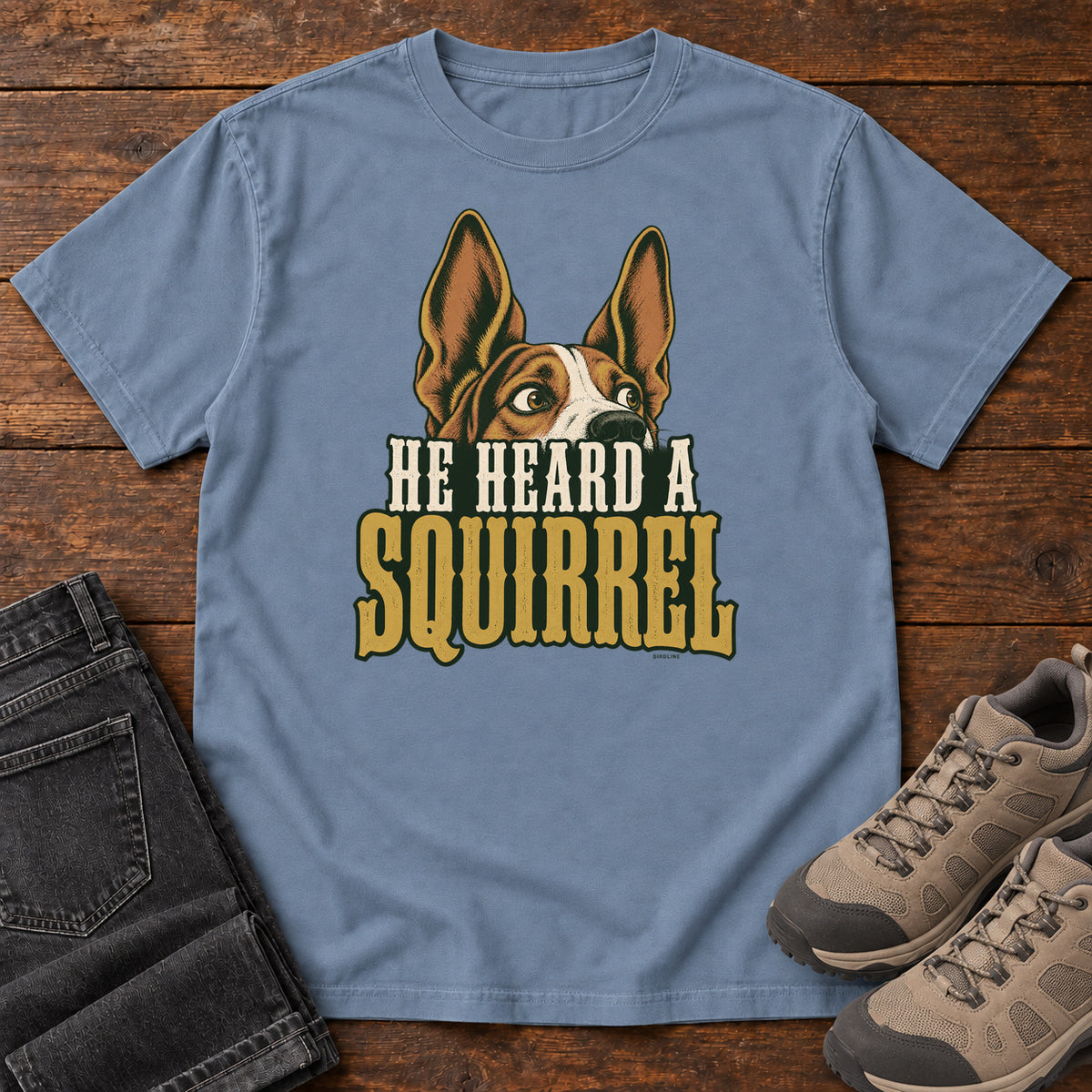 Blue Jean He Heard a Squirrel T-Shirt with an alert dog with upright ears above He Heard a Squirrel lettering; front flat lay on wood.