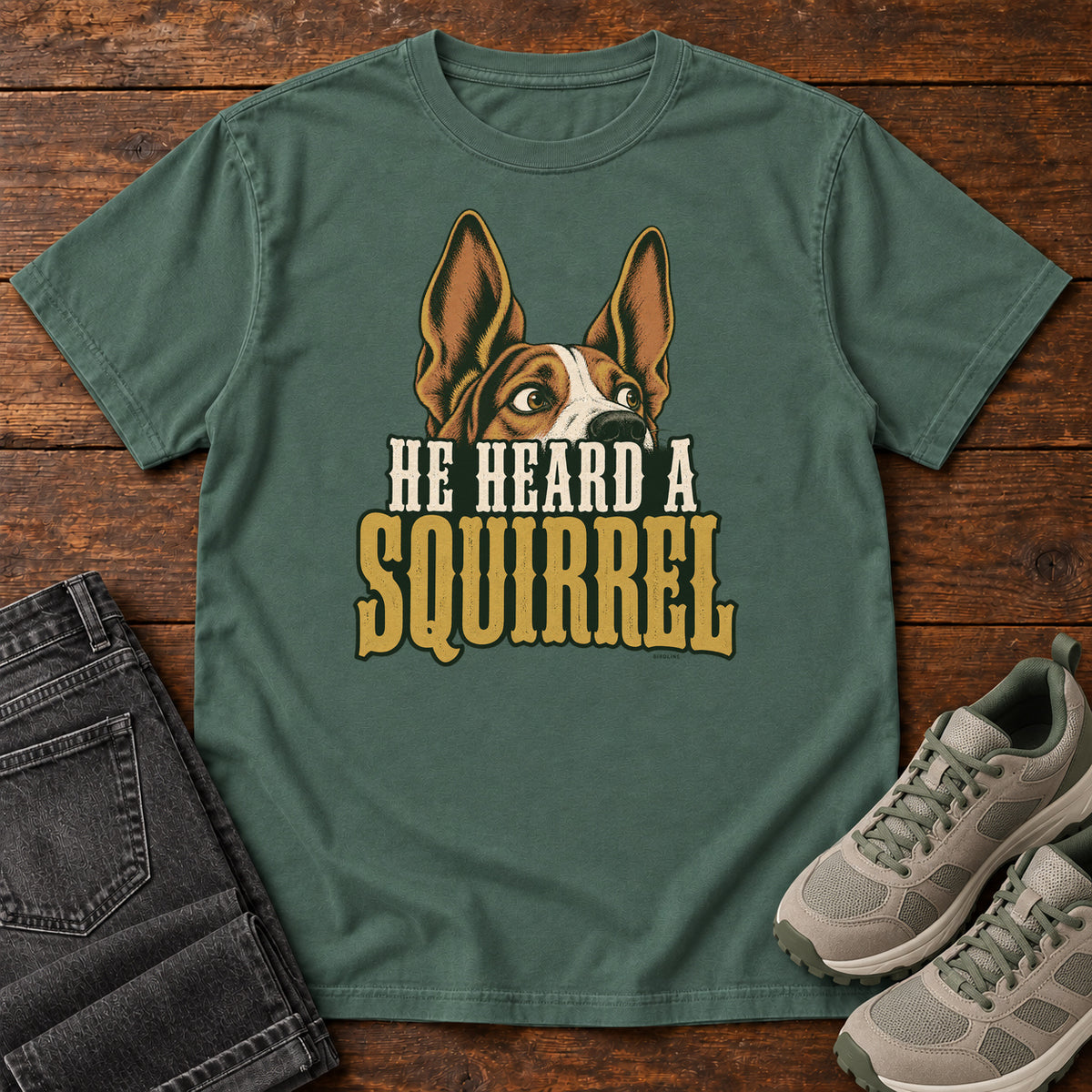Blue Spruce He Heard a Squirrel T-Shirt with an alert dog with upright ears above He Heard a Squirrel lettering; front flat lay on wood.