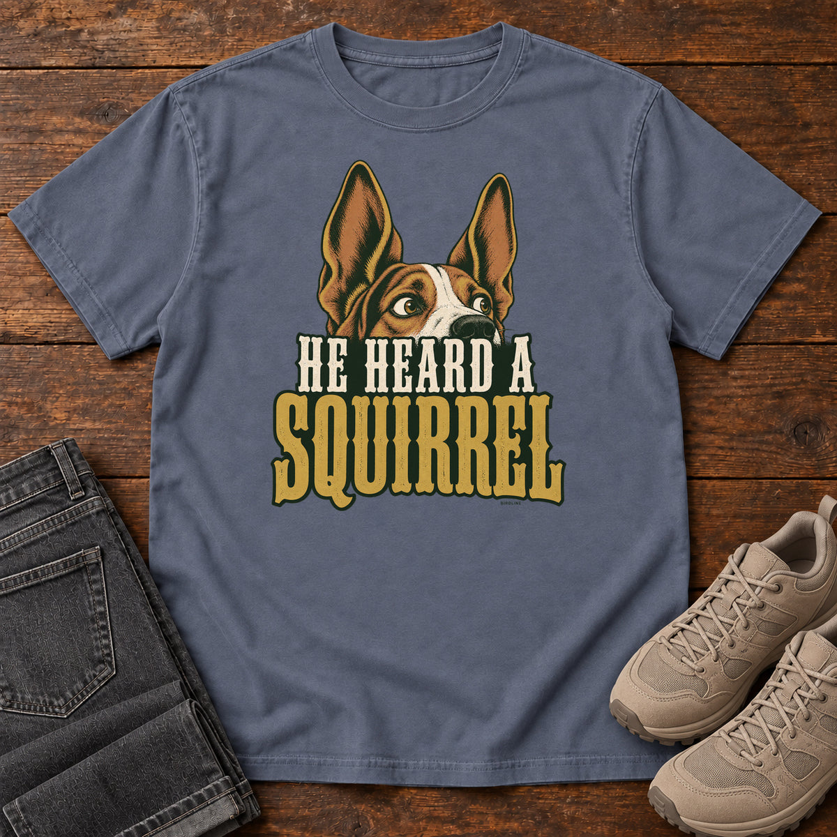 Denim He Heard a Squirrel T-Shirt with an alert dog with upright ears above He Heard a Squirrel lettering; front flat lay on wood.