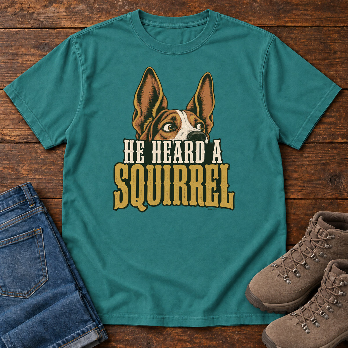 Emerald He Heard a Squirrel T-Shirt with an alert dog with upright ears above He Heard a Squirrel lettering; front flat lay on wood.